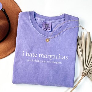 May include: A violet short-sleeved t-shirt with a crew neckline and a Comfort Colors brand label. The shirt features white text that reads i hate margaritas just kidding, can you imagine? A thin gold-toned chain necklace rests on the fabric.