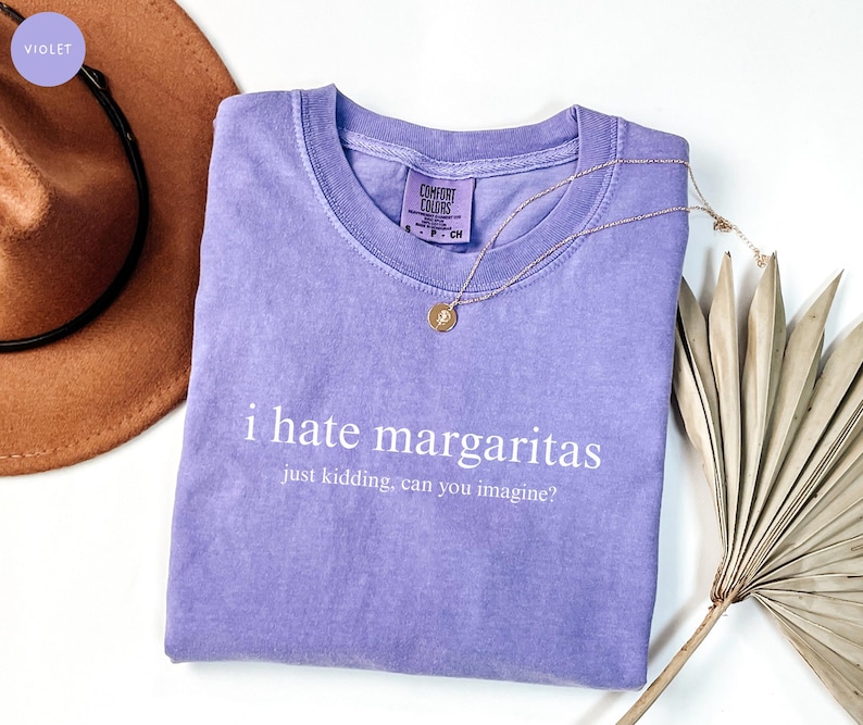 May include: A violet short-sleeved t-shirt with a crew neckline and a Comfort Colors brand label. The shirt features white text that reads i hate margaritas just kidding, can you imagine? A thin gold-toned chain necklace rests on the fabric.