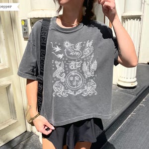 May include: A charcoal gray short-sleeve t-shirt featuring a white graphic print of multiple dragons and a central sun face. The shirt has a crew neckline and a relaxed fit. A black bag strap with the repeated white text BAGGU is visible.