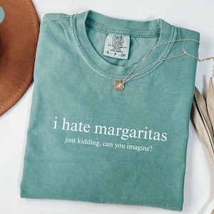 May include: A light green t-shirt with a crew neckline and a Comfort Colors brand label. The shirt features white text that reads: i hate margaritas, just kidding, can you imagine? A thin silver-toned chain necklace with a small round pendant rests on top.