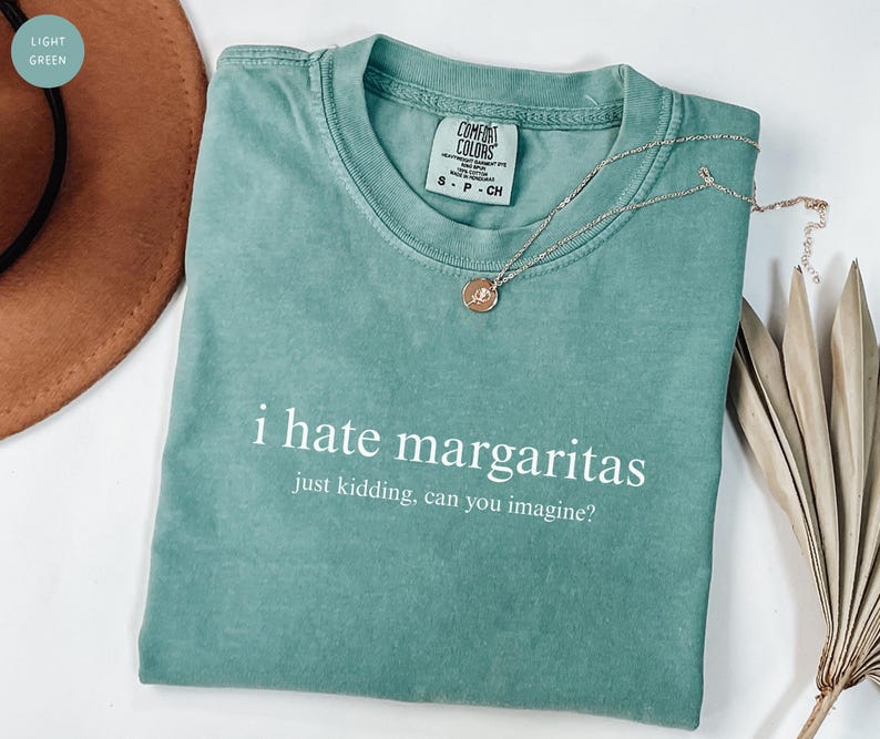 May include: A light green t-shirt with a crew neckline and a Comfort Colors brand label. The shirt features white text that reads: i hate margaritas, just kidding, can you imagine? A thin silver-toned chain necklace with a small round pendant rests on top.