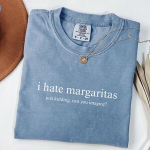 May include: A blue jean colored t-shirt with a crew neckline and the white printed text i hate margaritas just kidding, can you imagine? The shirt features a Comfort Colors brand label on the inside collar and is paired with a gold tone necklace.