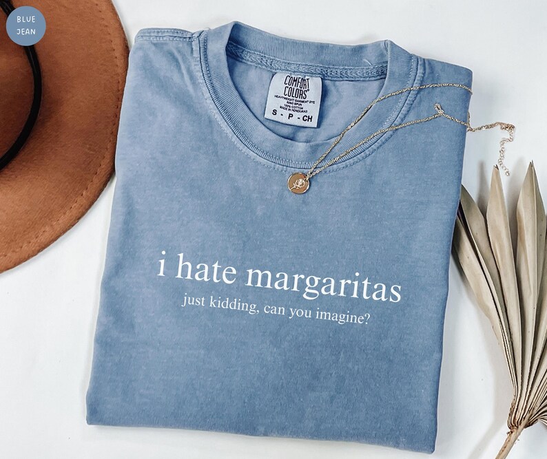 May include: A blue jean colored t-shirt with a crew neckline and the white printed text i hate margaritas just kidding, can you imagine? The shirt features a Comfort Colors brand label on the inside collar and is paired with a gold tone necklace.