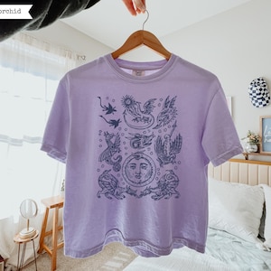 May include: A light purple short-sleeve t-shirt featuring a dark blue graphic print of various mythical dragons and a central sun face. The shirt has a crew neckline and is displayed on a wooden hanger. The word orchid is printed in the corner.