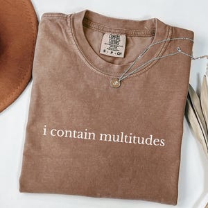Op de afbeelding: Een bruin Comfort Colors T-shirt met de witte tekst "i contain multitudes". Een zilveren ketting met een hanger ligt op het shirt. Een bruine hoed en een gedroogde plant staan op de achtergrond.