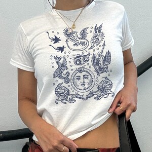 May include: White t-shirt featuring a dark blue illustration of mythical creatures and a sun with a face. The short-sleeved shirt has a crew neck and is made of a soft material. The design includes dragons and other fantasy elements.