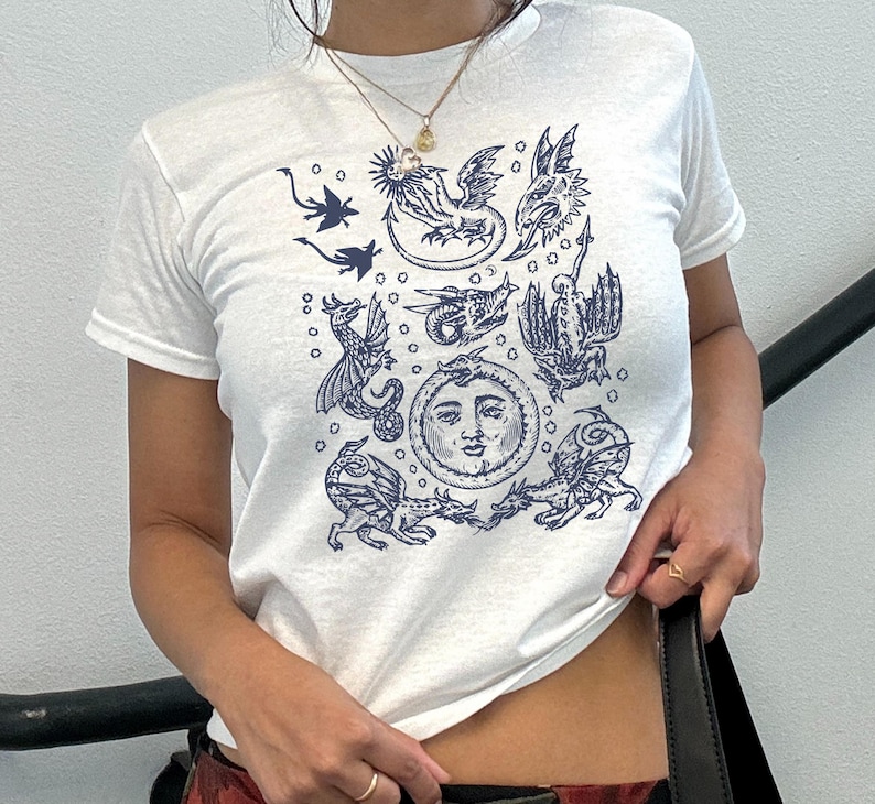 May include: White t-shirt featuring a dark blue illustration of mythical creatures and a sun with a face. The short-sleeved shirt has a crew neck and is made of a soft material. The design includes dragons and other fantasy elements.
