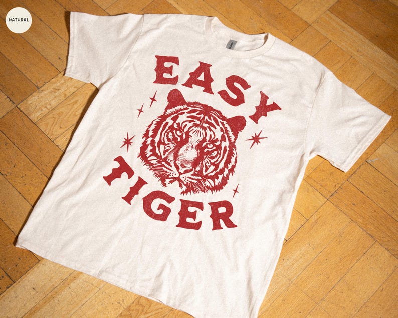 May include: Cream-colored t-shirt with a red tiger graphic and the words "EASY TIGER". The design includes a tiger's face and starburst accents. The shirt is made of a soft material.