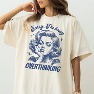 May include: Cream-colored t-shirt featuring a blue illustration of a woman with the text "Sorry, I'm busy OVERTHINKING". The graphic design has a vintage aesthetic. The shirt is a casual, oversized fit.