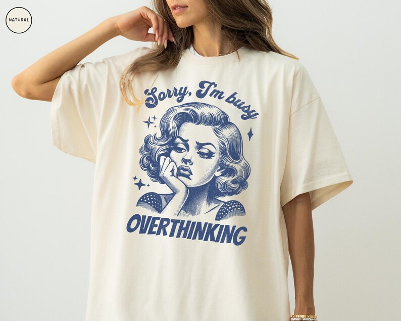 May include: Cream-colored t-shirt featuring a blue illustration of a woman with the text "Sorry, I'm busy OVERTHINKING". The graphic design has a vintage aesthetic. The shirt is a casual, oversized fit.