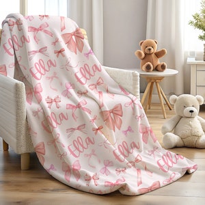 Pode incluir: Um cobertor branco com um padr&atilde;o de la&ccedil;os rosa e o nome "Ella" em escrita rosa. O cobertor est&aacute; sobre uma cadeira. Os la&ccedil;os variam em tamanho e tom, criando uma est&eacute;tica suave e feminina. Um urso de pel&uacute;cia est&aacute; por perto.