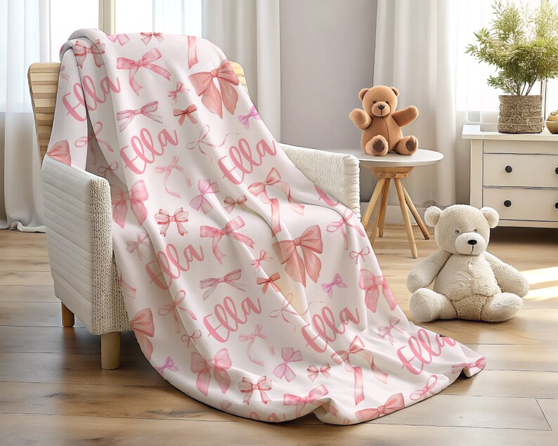 Pode incluir: Um cobertor branco com um padr&atilde;o de la&ccedil;os rosa e o nome "Ella" em escrita rosa. O cobertor est&aacute; sobre uma cadeira. Os la&ccedil;os variam em tamanho e tom, criando uma est&eacute;tica suave e feminina. Um urso de pel&uacute;cia est&aacute; por perto.
