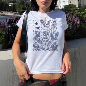 May include: White t-shirt with a dark blue illustration featuring mythical creatures, a sun, and other celestial elements. The short-sleeved shirt is made of a soft material.