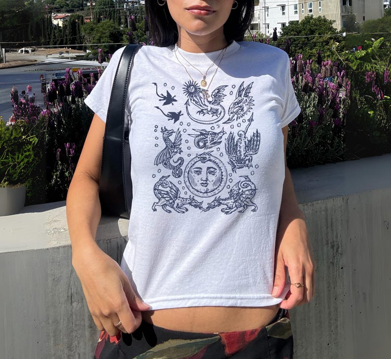 May include: White t-shirt with a dark blue illustration featuring mythical creatures, a sun, and other celestial elements. The short-sleeved shirt is made of a soft material.
