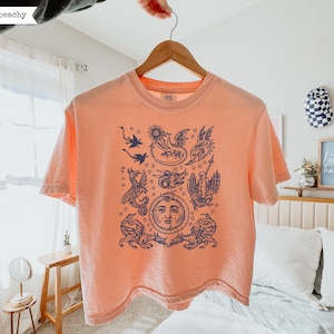 May include: A peach-colored short-sleeve t-shirt featuring a dark blue graphic print on the front. The design includes multiple illustrations of winged dragons and a central sun face surrounded by small circular accents in a detailed ink-style drawing.
