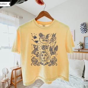 May include: A butter yellow short-sleeve t-shirt featuring a dark blue graphic print of various dragons and a central sun face. The design includes multiple mythical creatures in a detailed, etched style arranged around a circular sun illustration.