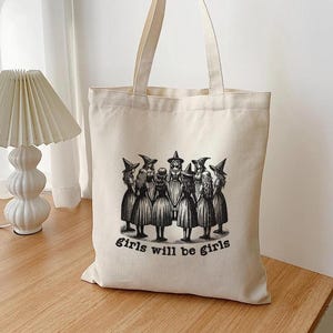 May include: A white canvas tote bag with a black and white illustration of a group of witches in a circle. The text "girls will be girls" is printed below the illustration.