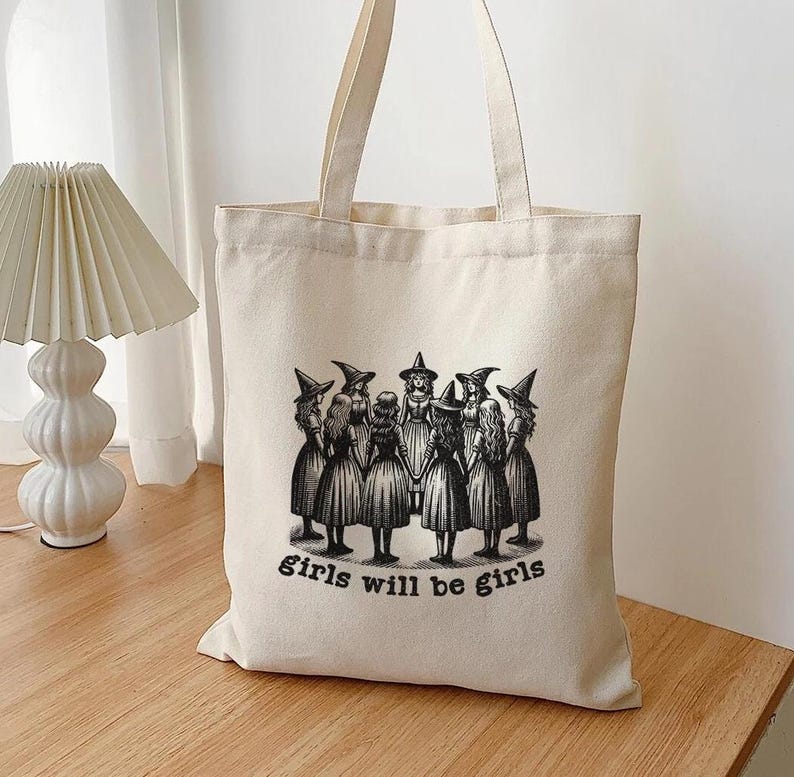 May include: A white canvas tote bag with a black and white illustration of a group of witches in a circle. The text "girls will be girls" is printed below the illustration.