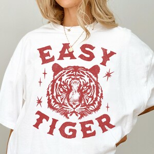 May include: White t-shirt with a red graphic design. The design features the text "EASY TIGER" above and below a detailed tiger head illustration. The shirt has a relaxed fit and short sleeves.