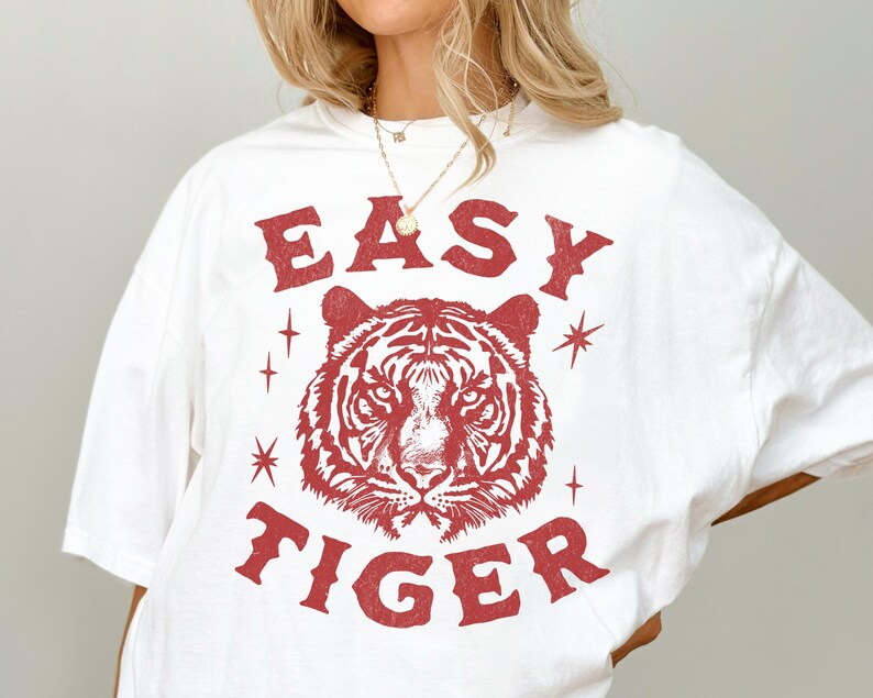 May include: White t-shirt with a red graphic design. The design features the text "EASY TIGER" above and below a detailed tiger head illustration. The shirt has a relaxed fit and short sleeves.