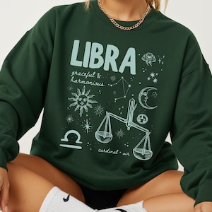 May include: A dark green long-sleeve sweatshirt featuring a light blue graphic print. The design includes the text LIBRA, graceful & harmonious, and cardinal air, alongside illustrations of a sun, moon, stars, and a set of scales.