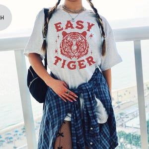 May include: Ash-colored t-shirt with a red tiger graphic and the words "EASY TIGER". The shirt is a light gray color and has a relaxed fit. The tiger graphic is centered on the front of the shirt.