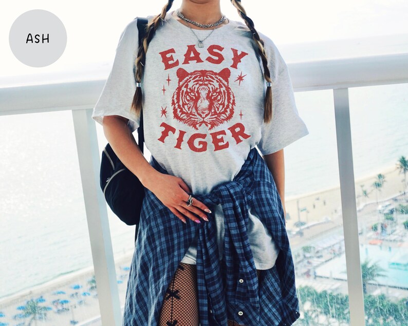 May include: Ash-colored t-shirt with a red tiger graphic and the words "EASY TIGER". The shirt is a light gray color and has a relaxed fit. The tiger graphic is centered on the front of the shirt.