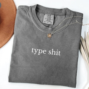 Op de afbeelding: Een grijze t-shirt met de woorden "type shit" in het wit gedrukt. Een zilveren ketting met een hanger ligt op de shirt. Een bruine hoed en een gedroogde plant staan op de achtergrond.