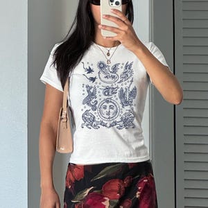 May include: White t-shirt with a navy blue illustration of mythical creatures and a sun face. The shirt is short-sleeved and worn with a floral skirt and a light brown purse. The person is wearing sunglasses and a necklace.