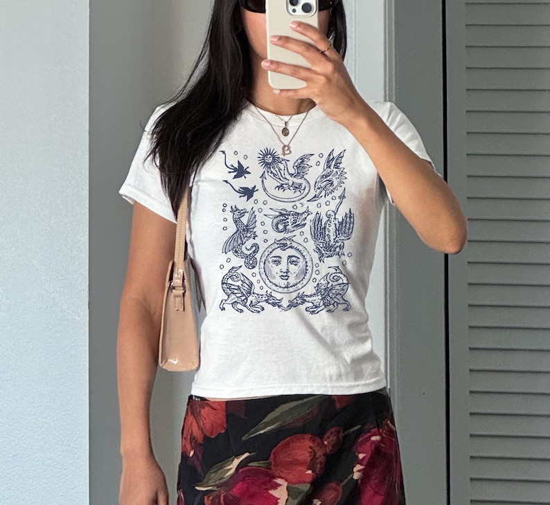 May include: White t-shirt with a navy blue illustration of mythical creatures and a sun face. The shirt is short-sleeved and worn with a floral skirt and a light brown purse. The person is wearing sunglasses and a necklace.
