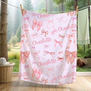 Pode incluir: Um cobertor de beb&ecirc; branco com um padr&atilde;o de la&ccedil;os rosa em aquarela e o nome "Charlotte" em escrita rosa. O cobertor est&aacute; pendurado em um varal ao ar livre, com uma floresta ao fundo.