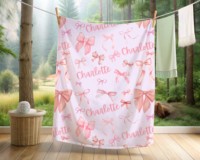 Pode incluir: Um cobertor de beb&ecirc; branco com um padr&atilde;o de la&ccedil;os rosa em aquarela e o nome "Charlotte" em escrita rosa. O cobertor est&aacute; pendurado em um varal ao ar livre, com uma floresta ao fundo.