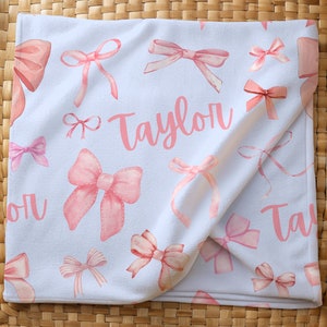 Pode incluir: Cobertor de beb&ecirc; branco com um padr&atilde;o de la&ccedil;os rosa em aquarela. O nome "Taylor" est&aacute; escrito em rosa no cobertor. O cobertor est&aacute; dobrado e colocado em uma cesta de vime.