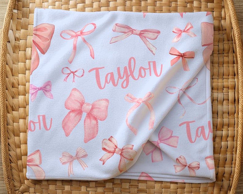 Pode incluir: Cobertor de beb&ecirc; branco com um padr&atilde;o de la&ccedil;os rosa em aquarela. O nome "Taylor" est&aacute; escrito em rosa no cobertor. O cobertor est&aacute; dobrado e colocado em uma cesta de vime.
