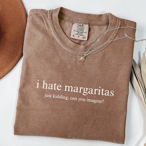 May include: A brown t-shirt with the phrase "i hate margaritas, just kidding, can you imagine?" printed in white. The shirt has a crew neck and a Comfort Colors label. A silver necklace with a pendant is on the shirt.