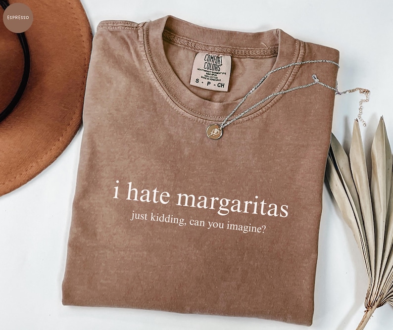 May include: A brown short-sleeved t-shirt with a crew neckline. The shirt features white text that reads "i hate margaritas" followed by "just kidding, can you imagine?" The garment has a Comfort Colors brand label on the inside collar.