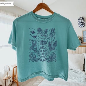 May include: A chalky mint short-sleeve t-shirt featuring a dark blue graphic print of various mythical dragons and a central sun face. The design includes multiple winged creatures arranged in a grid-like pattern on the front of the garment.
