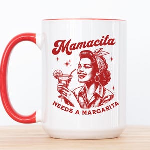 Puede incluir: Taza de cerámica blanca con borde y asa rojos. La taza presenta una ilustración roja de una mujer con una margarita y el texto "Mamacita Needs a Margarita".
