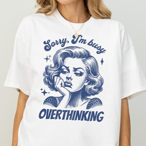 May include: White t-shirt with a blue graphic of a woman with the text "Sorry, I'm busy OVERTHINKING". The illustration features a vintage style with stars and a thoughtful pose.