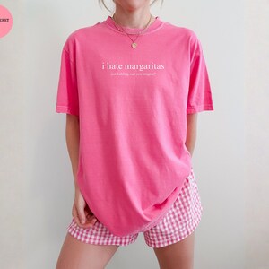 May include: A bright pink short-sleeved t-shirt featuring the white text "i hate margaritas" followed by the smaller text "just kidding, can you imagine?". The shirt is paired with pink and white gingham patterned shorts and a gold necklace.