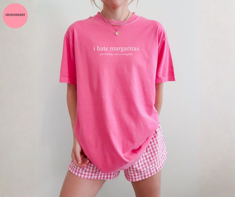 May include: A bright pink short-sleeved t-shirt featuring the white text "i hate margaritas" followed by the smaller text "just kidding, can you imagine?". The shirt is paired with pink and white gingham patterned shorts and a gold necklace.