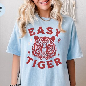 May include: Light blue t-shirt with the text "EASY TIGER" in red, featuring a tiger head graphic. The shirt has short sleeves and a relaxed fit. The words "LIGHT BLUE" are in a light blue circle in the upper left corner.