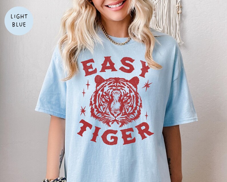 May include: Light blue t-shirt with the text "EASY TIGER" in red, featuring a tiger head graphic. The shirt has short sleeves and a relaxed fit. The words "LIGHT BLUE" are in a light blue circle in the upper left corner.
