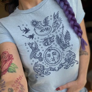 May include: Light blue t-shirt featuring a dark blue dragon graphic. The design includes various dragon illustrations, a sun, and a moon with a face. The shirt is short-sleeved and made of a soft material.