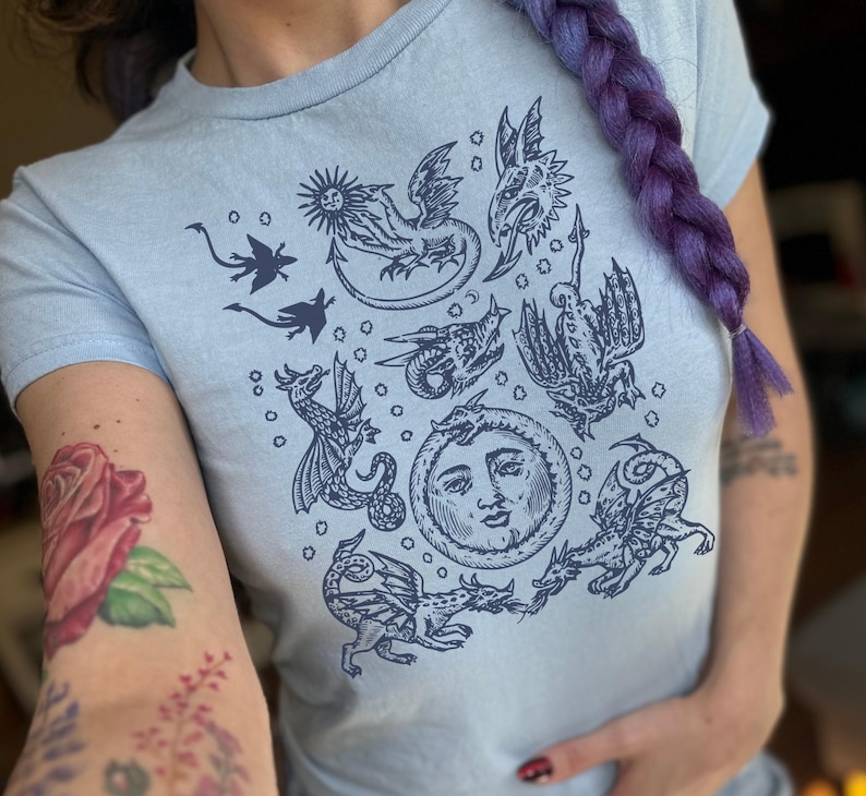 May include: Light blue t-shirt featuring a dark blue dragon graphic. The design includes various dragon illustrations, a sun, and a moon with a face. The shirt is short-sleeved and made of a soft material.