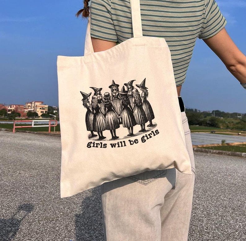 May include: A white tote bag with a black and white illustration of a group of witches. The text "girls will be girls" is printed on the bag.