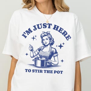 以下が含まれることがあります： 青いグラフィックの白いTシャツ。グラフィックは、鍋をかき混ぜる女性のヴィンテージイラストで、「I'M JUST HERE TO STIR THE POT」というテキストが一致する青いフォントで表示されています。シャツは半袖です。