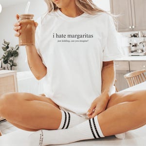 May include: A white short-sleeved t-shirt featuring the black printed text 'i hate margaritas' followed by the smaller text 'just kidding, can you imagine?' centered on the chest. The shirt has a crew neckline and a relaxed, loose-fitting silhouette.