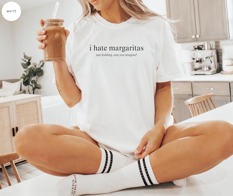 May include: A white short-sleeved t-shirt featuring the black printed text 'i hate margaritas' followed by the smaller text 'just kidding, can you imagine?' centered on the chest. The shirt has a crew neckline and a relaxed, loose-fitting silhouette.