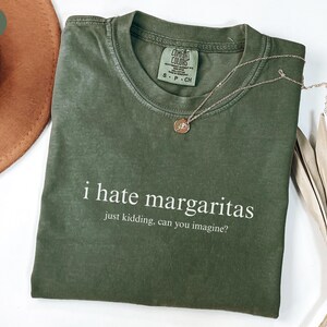 May include: A sage green short-sleeve t-shirt with a crew neckline. The shirt features white text that reads: i hate margaritas, just kidding, can you imagine? A thin gold-tone chain necklace with a small circular pendant rests on the collar.
